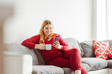 Smiling plus size young woman sitting on sofa in living room at home, holding mobile phone and cup
