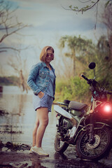 beautiful woman wearing blue jeans jacket standing beside enduro motorcycle against colorful natural  background