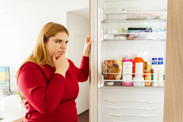 Beautiful body positive woman opened refrigerator, choosing food in kitchen in cozy apartment