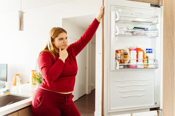 Pensive, attractive Caucasian plus size woman opened refrigerator, choosing food in kitchen