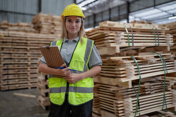 Portrait of caucasian female worker standing in warehouse facility holds clipboard looking at camera smiling. Woman engineer employee wear safety hardhat working in wooden pallet production factory.