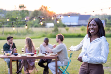 A joyful gathering on a sunny day encapsulates a beautiful young black woman in a white blouse, laughing and savoring a garden barbecue amidst friends, wine, and outdoor picnic fun. The essence of
