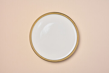 Empty round ceramic white plate with a gilded edge on a beige background, top view
