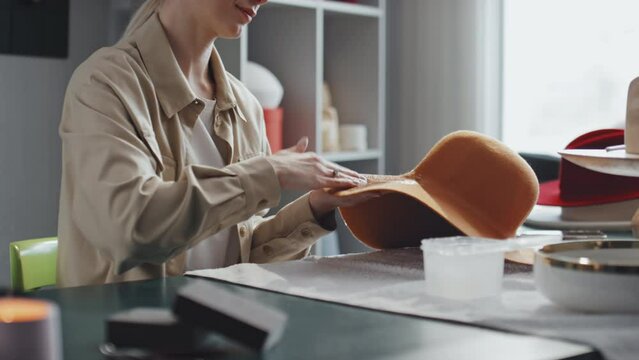 Midsection shot of professional female hatter sitting in her workshop coating orange felt hat with stiffener