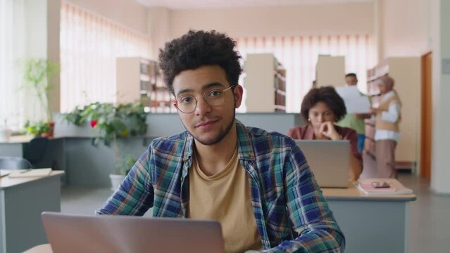 Waist up slowmo portrait of Biracial guy wearing glasses and blue checkered shirt sitting at desk in library with laptop, studying language together with other migrants