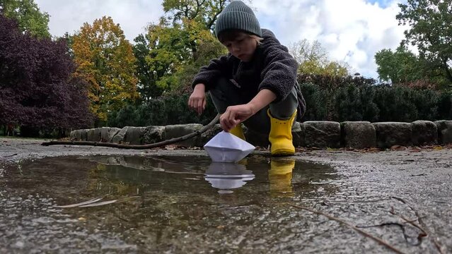 Child Sits by a Puddle, Playing with Paper Boats on a Cold Autumn Day. High quality 4k footage.