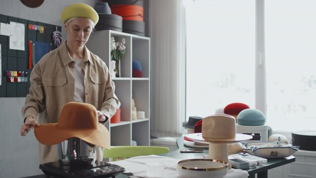 Medium shot of Caucasian female milliner putting orange felt hat on stockpot with boiling water