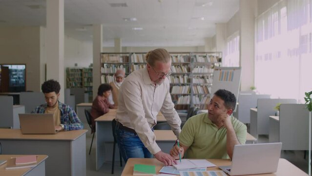 Medium shot of mid aged Caucasian male English teacher helping Middle Eastern migrant student to do grammar worksheet during group lesson in modern libraryMedium shot of mid aged Caucasian male Englis