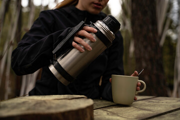 exterior of a Glamping , close-up detail Girl uses thermos of coffee on a wooden table outdoors during autumn. © Alberto Cotilla