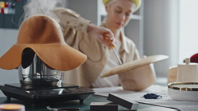 Medium close-up selective focus shot of steaming stockpot with felt hat on while female milliner making new wooden mannequin