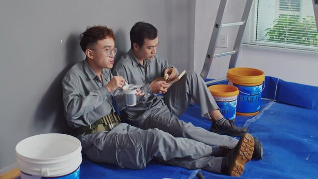 Long shot of two repairmen in uniform having lunch after painting walls at construction site