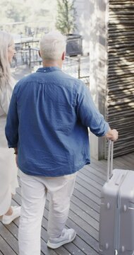Vertical video of happy senior caucasian couple walking with suitcase, slow motion