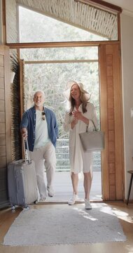 Vertical video of happy senior caucasian couple walking through door with suitcase, slow motion