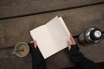 woman's hands reading a book and drinking coffee on a wooden table in the open air.