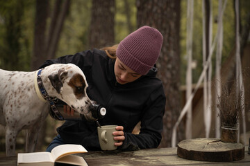 Girl in winter hat is sitting while drinking coffee with her dog outdoors.