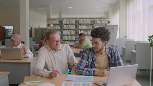 Medium shot of Biracial migrant student listening to adult Caucasian male tutor explaining him English grammar in modern library