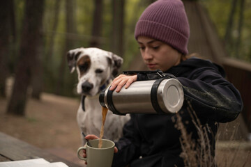 Girl in winter hat is sitting while pouring coffee in the cup next to her dog are outdoors.