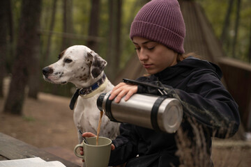 Girl in winter hat is sitting while pouring coffee in the cup next to her dog are outdoors.