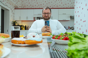 Obraz premium Man with extra weight photographing food on lunchtime at home in the kitchen. Concept of dieting and loosing weight, fatman only picturing food but not eat it