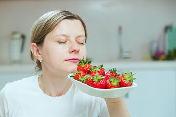 Woman smelling ripe red strawberries on white plate. Concept of eating natural sugar products and stay healthy and thin. Dieting concept, raw fruits diet