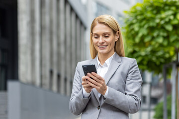 Successful woman boss walks outside office building outdoors, in business suit, businesswoman uses app on phone, smiles contentedly reads internet pages, browses news, writes text message, call