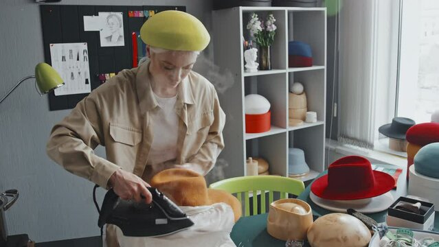 Medium shot of Caucasian female hatter wearing yellow beret and beige raincoat ironing felt hat using cheesecloth