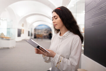 Side view of young caucasian woman student visiting museum exhibition and reading brochure. Light...