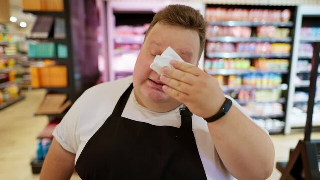 A tired supermarket worker, Overweight man, in a white T-shirt and a black apron wipes sweat from his forehead with a white napkin. Taking a break during a hard day at work