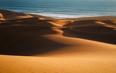 Curvy dunes of Namib Desert in front of calm ocean waves at sunset, south of Walvis Bay, in the...