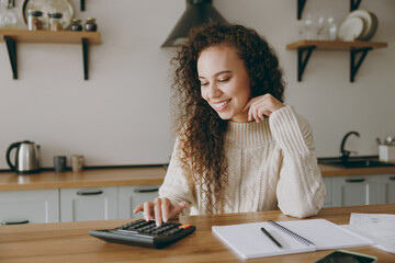 Young housewife woman of African American ethnicity wears casual clothes sweater use calculator studying prepare for exam sit at table in light kitchen at home alone. Lifestyle cooking food concept.
