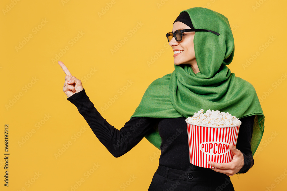 Wall mural Young muslim woman in 3d glasses wears green hijab abaya black clothes watch movie film hold bucket of popcorn in cinema point aside isolated on plain yellow background. Uae islam religious concept.