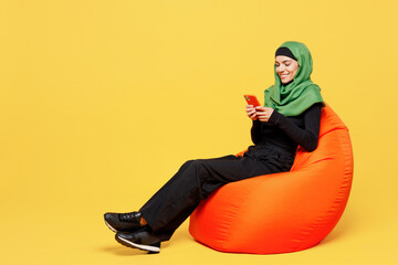 Full body young happy arabian asian muslim woman wears green hijab abaya black clothes sit in bag chair use mobile cell phone isolated on plain yellow background. People uae islam religious concept.