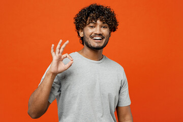 Young smiling cheerful fun satisfied happy Indian man he wears t-shirt casual clothes showing okay ok gesture loking camera isolated on orange red color background studio portrait. Lifestyle concept.