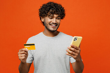 Young smiling happy Indian man he wears t-shirt casual clothes using mobile cell phone hold credit...
