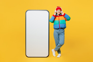 Full body young woman wear padded windbreaker jacket red hat casual clothes big huge blank screen mobile cell phone smartphone with area listen music in headphones isolated on plain yellow background