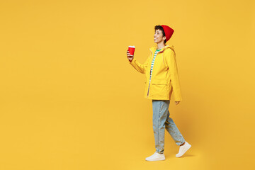 Full body young woman wears waterproof raincoat outerwear red hat hold takeaway paper cup coffee to go isolated on plain yellow background studio. Outdoors lifestyle wet fall weather season concept.