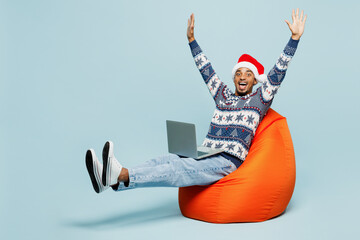 Full body young man wear knitted sweater Santa hat posing sit in bag chair hold use work on laptop pc computer isolated on plain pastel blue background. Happy New Year 2024 Christmas holiday concept.