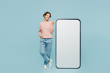 Full body young woman wear beige knitted sweater casual clothes point index finger on big huge blank screen mobile cell phone smartphone with workspace area isolated on plain pastel blue background.