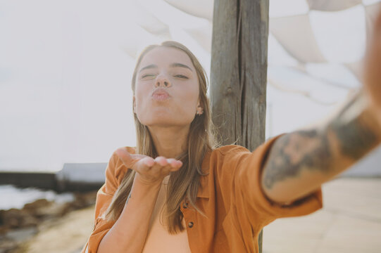 Close up young woman she wearing orange shirt beige top casual clothes doing selfie shot pov on mobile cell phone send air kiss rest relax walk outside in summer day. Urban lifestyle leisure concept.