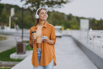 Young minded woman wear orange shirt casual clothes listen music in headphones use mobile cell phone drink coffee rest relax walk in spring city park outside in summer day. Urban lifestyle concept.