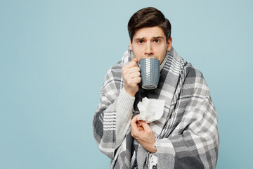 Young sad ill sick man wrapped in gray plaid drinking warm tea holding napkin isolated on plain blue background studio portrait. Healthy lifestyle disease virus treatment cold season recovery concept.