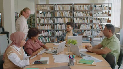 Medium shot of mature Caucasian male teacher handing clipboards with worksheets to diverse migrant students studying English language in group at library - Powered by Adobe