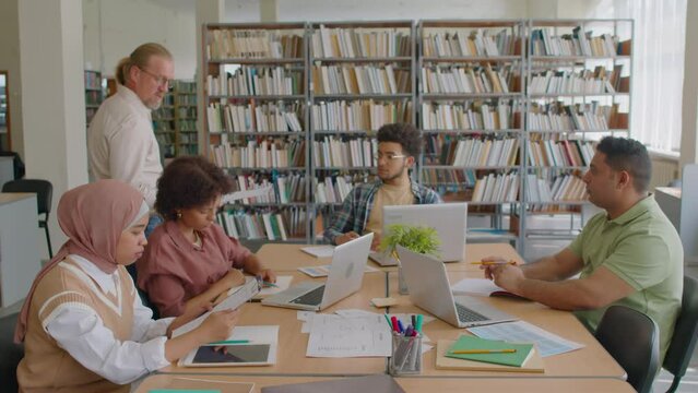 Medium shot of mature Caucasian male teacher handing clipboards with worksheets to diverse migrant students studying English language in group at library