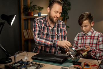 Father with son using soldering iron for repairing laptop computer at home. Concept of maintenance...