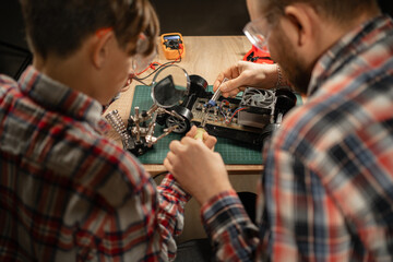 Father and son in safety glasses are soldering a broken machine on the control panel, view over the...