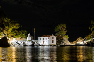 Enchanting Night View of Holy Mary Island in Parga Greece. White Chapel with Belfry Surrounded by Big Trees. Amazing Night Photography with the Sea that Reflects Lights.