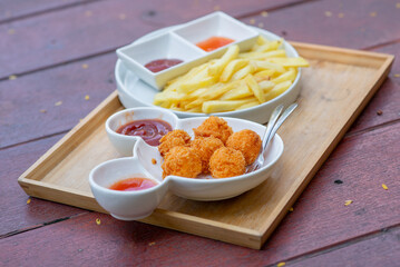 French fries and cheese balls on a white plate on a tray and a wooden board background Contains ketchup and chili sauce