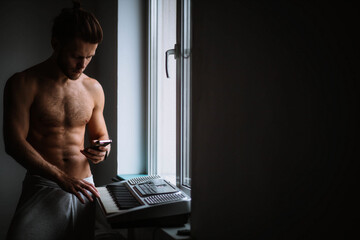sexy man with a naked torso at the window with a smartphone and a piano synthesizer. Online music lessons.