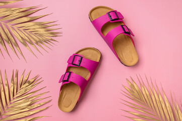 Fashionable leather bright pink birkenstock sandals, golden palm leaf on pink background, top view flat lay. Minimalistic concept of summer unisex shoes, flip-flop sandals for vacation, beach