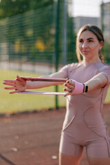 Young woman warming up and strengthening her arms with fitness band on the sports ground.